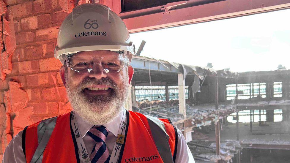 James Petter who has a bushy white beard and is wearing clear eye protector goggles, a white hard hat with Colemans written it and a hi-vis orange waistcoat over a white shirt and pink and blue tie. He is smiling broadly. Beyond him is a window and through it can be seen the part-demolished three story M&S in Northampton.