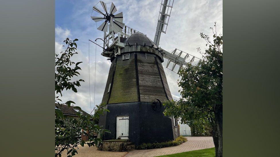 Fulbourn windmill, an early 19th Century building. It has a black base and above its white door is weathered looking boarding which rises to a conical top. Its white sails are on the far side of the mill. Its white fantail is on this side of the mill. It is surrounded by block paving and a house can be glimpsed on the left.
