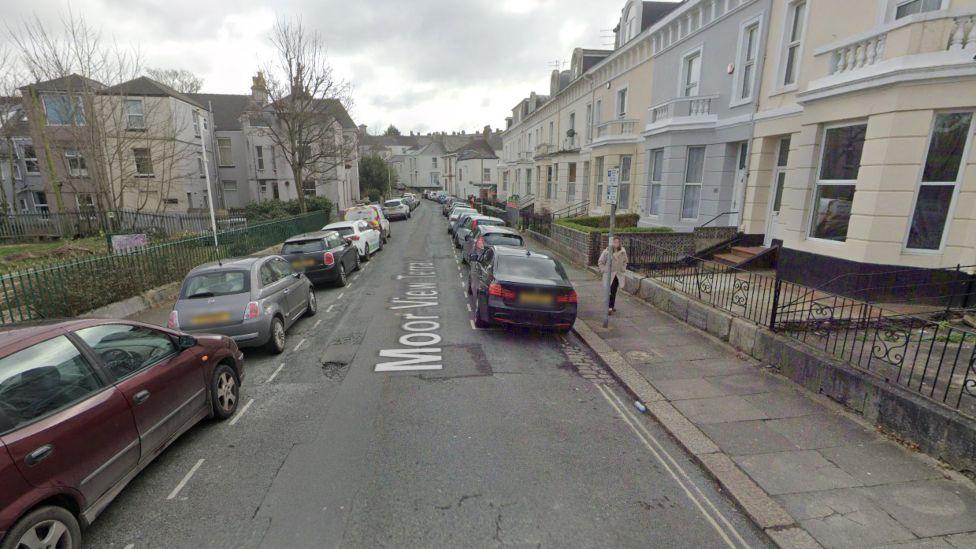 Google Maps image of Moor View Terrace. Cream and grey buildings are on the right-hand side of a narrow side street. A number of cars are parked on either side of the road.