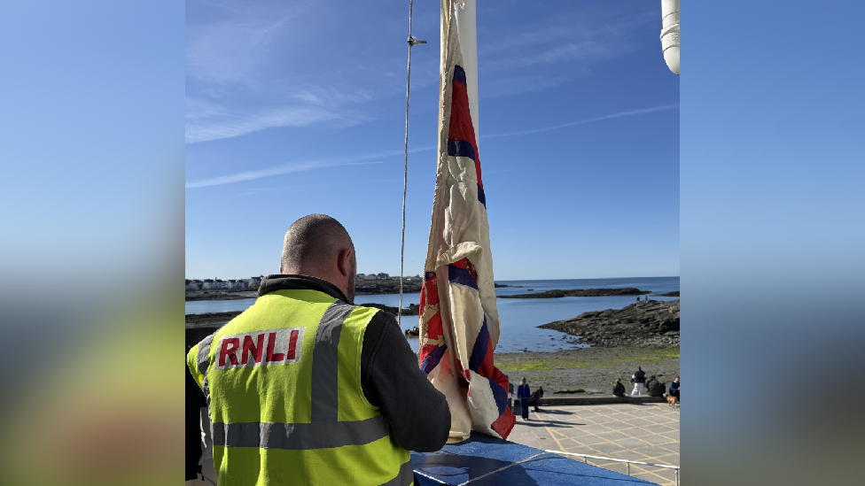 A RNLI volunteer putting the old flag on the flagpole. The man is facing the sea, away from the camera and is wearing a high vis yellow top with red RNLI writing on. It is a sunny day. 