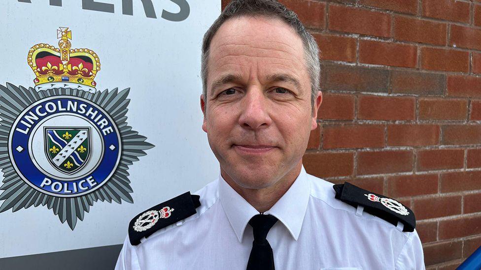 Chief Constable Paul Gibson with short dark hair he is wearing a white uniform shirt with epaulettes and is stood in front of a brick wall which has the forces badge on it