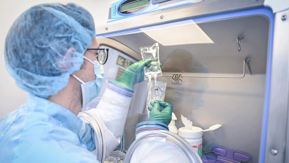 A scientist in the pharmacy clinical trials unit wearing a face mask and hair net is inserting a needle into a bag of fluid.