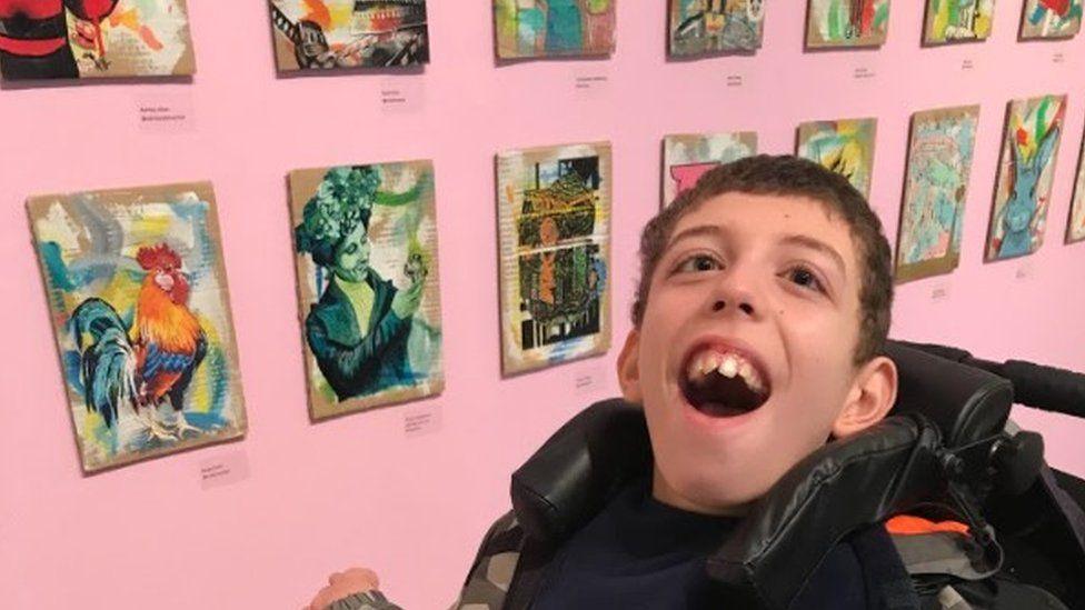 Noah, who is sat in a wheelchair, smiling at the camera. He is in front of the cardboard paintings that are on display on a pink wall.