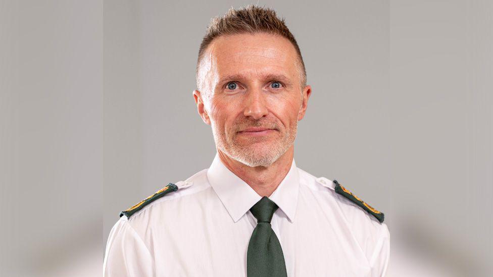 Jason Killens looking at the camera smiling. He has short cropped hair and is wearing a white shirt with a green tie and green and gold embossed ambulance shoulder badges.