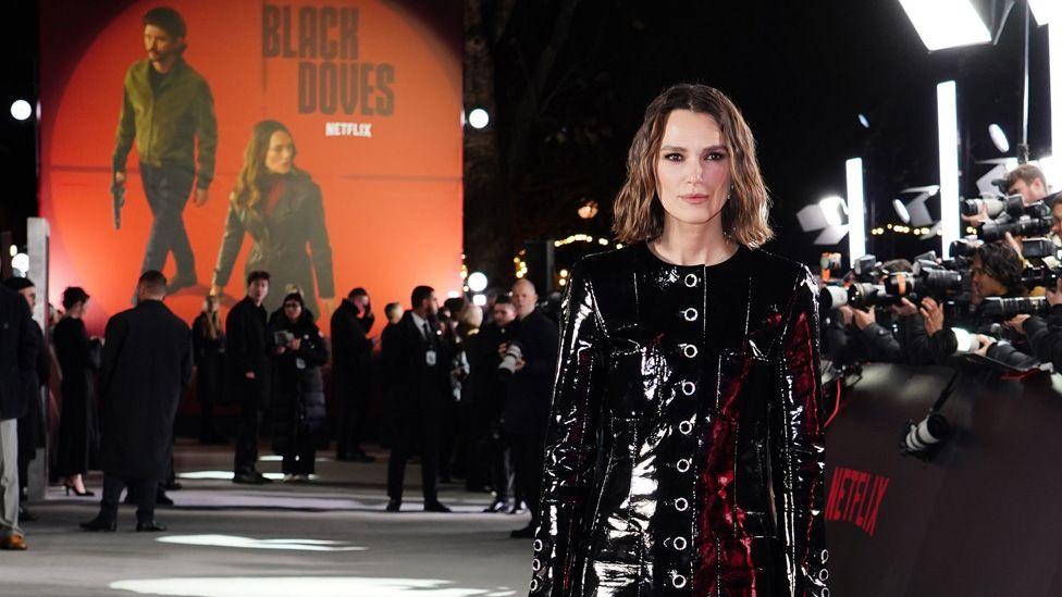 Keira Knightley in an all-black outfit with photographers and a large promotional image on a wall at the premiere of Black Doves in London on 3 December