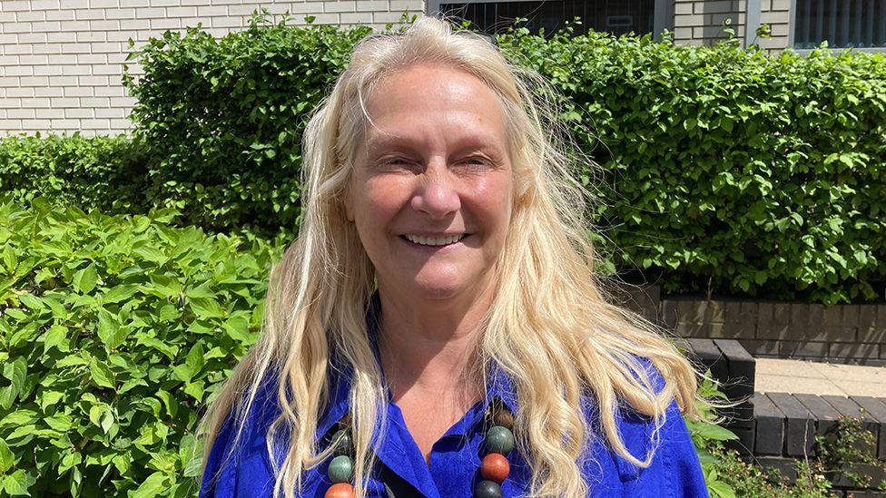 Head-and-shoulders image of Liz Crook standing outside in the sunshine in front of a green hedge. She has long blonde hair and is wearing a bright blue blouse and a long, multi-coloured chunky-beaded necklace.