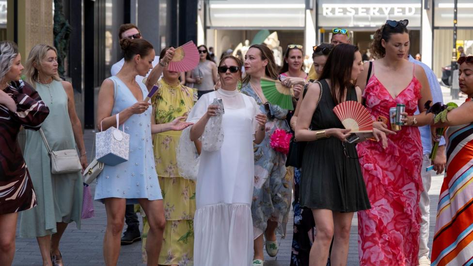 A bride walks through the centre of London with female guests in brightly coloured dresses surround her trying to fan her to keep cool