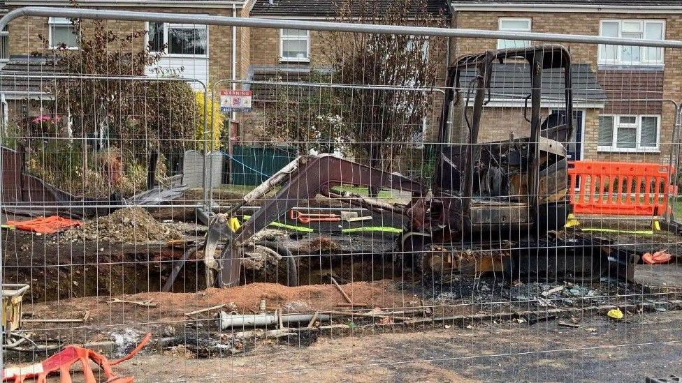 A metal fence surrounds a digger which has been burnt. There are houses in the background and several trees.