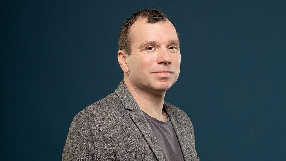 A headshot of a man with short brown hair, turned slightly to the side as he stands in front of a dark blue background. He's wearing a dark grey t-shirt under a lighter grey suit jacket.