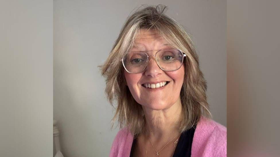 A woman with blonde hair and glasses, wearing a pink cardigan over a black top
