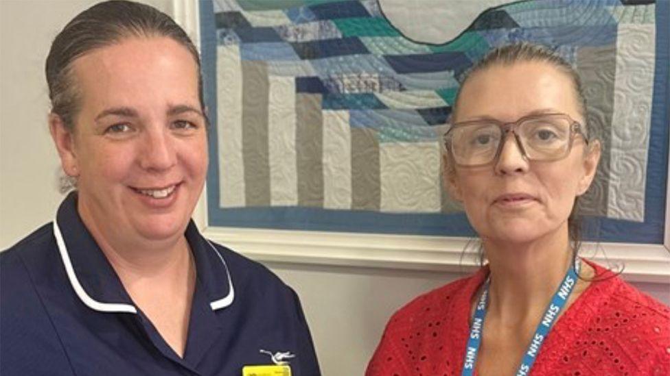 Vicki Lang in a navy nurses uniform with her brown hair tied back and Claire Hickson in a red lace top, glasses and NHS lanyard standing in front of an applique artwork in blue grey and white