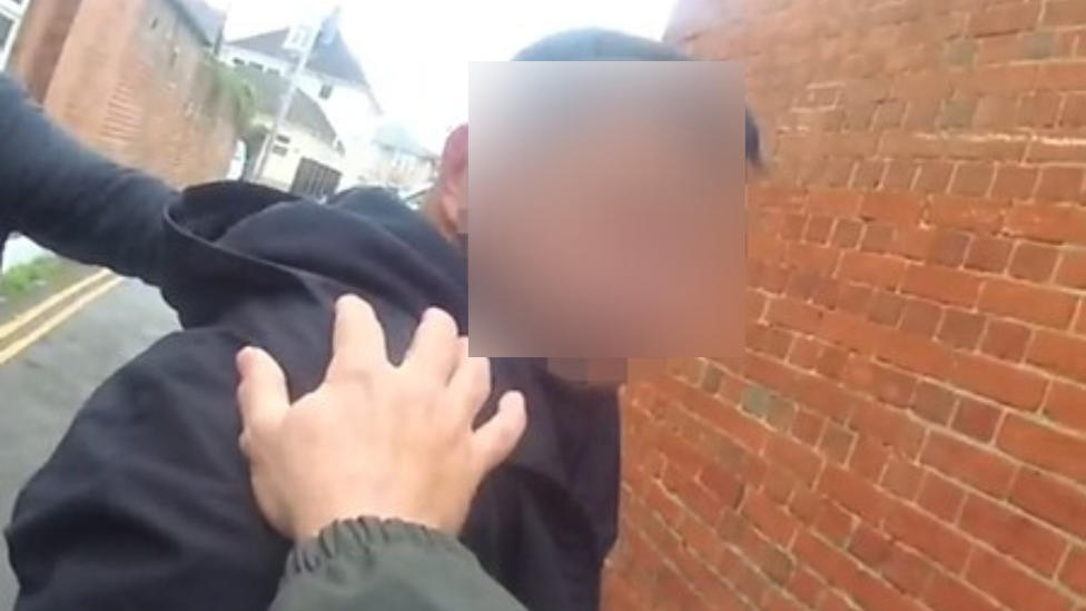 A man in a black hoodie seemingly being arrested has two hands from officers on his shoulders. His face is obscured by blurring.