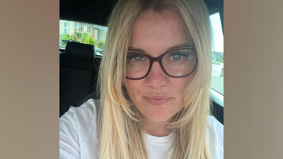 Becca looks at the camera in a selfie taken in a car. She has long blond hair and is wearing dark rimmed glasses and a white t-shirt.