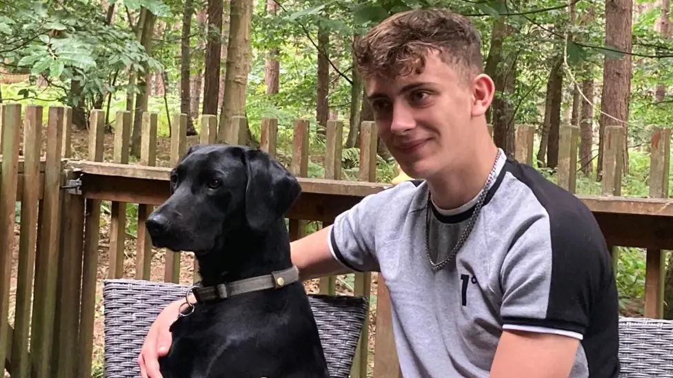 Image of Cory Chilvers sitting with his arms around a black Labrador dog. He has light brown hair and is wearing a grey T-shirt. He has a silver chain around his neck.