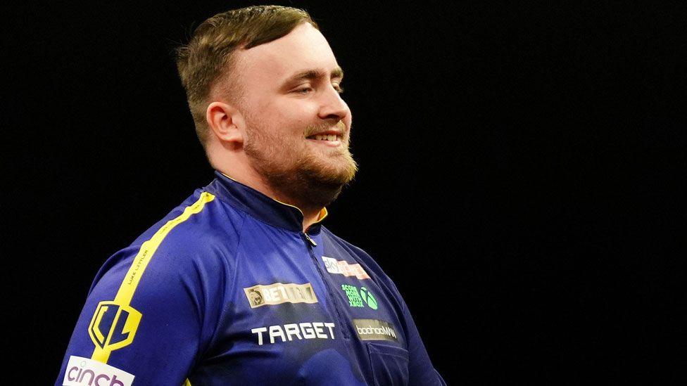 Luke Littler wearing a blue top covered in advertising slogans. He is sideways on, walking, and has short light brown hair and a light brown beard. He is smiling and taking part in BetMGM Premier League at P&J Live, Aberdeen, on 15 May.