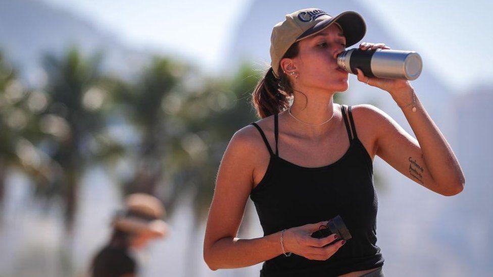 A woman drinks water on Copacabana Beach in Rio de Janeiro, Brazil, during a heatwave. She is wearing a black vest top and a light brown cap. Palm trees are visible in the background.