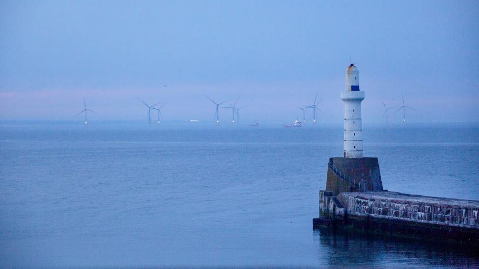 A white lighthouse, with wind turbines and offshore supply vessels in the distance in the sea off Aberdeen, under a blue/pink sky.