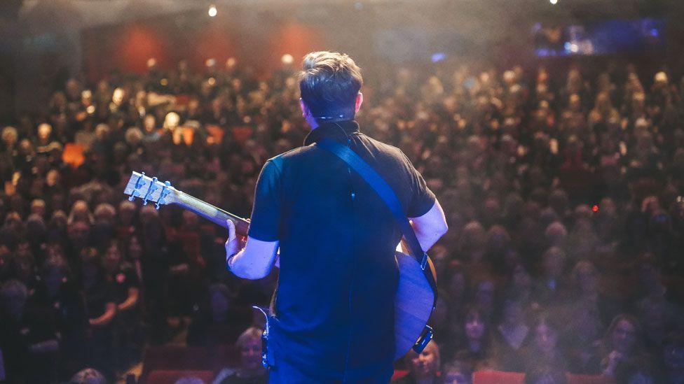 Back view of male guitarist wearing black and facing 1,200 singers, Westcliff-on-Sea