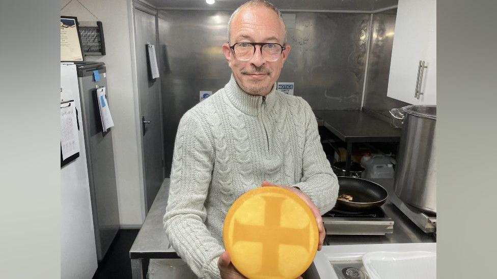 Matthew Lloyd inside his cheese factory. He is wearing a beige jumper with black glasses, and is holding a large, yellow wheel of cheese