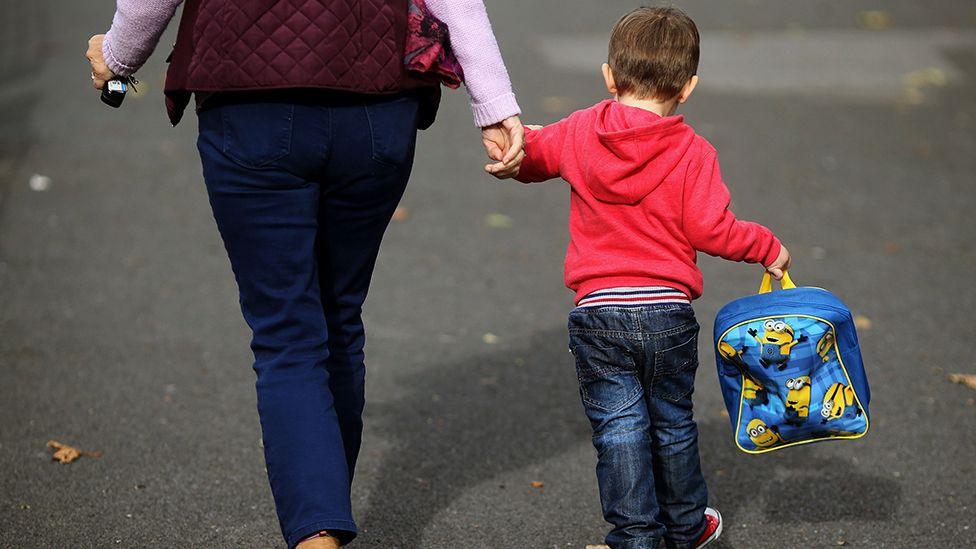 Generic image of a woman holding the hand of a small boy who is wearing jeans and a red hoodie and is holding a blue minions rucksack. They are walking away from the camera so their faces cannot be seen. The woman can be seen from the waist down and is wearing jeans and a dark red gillet.