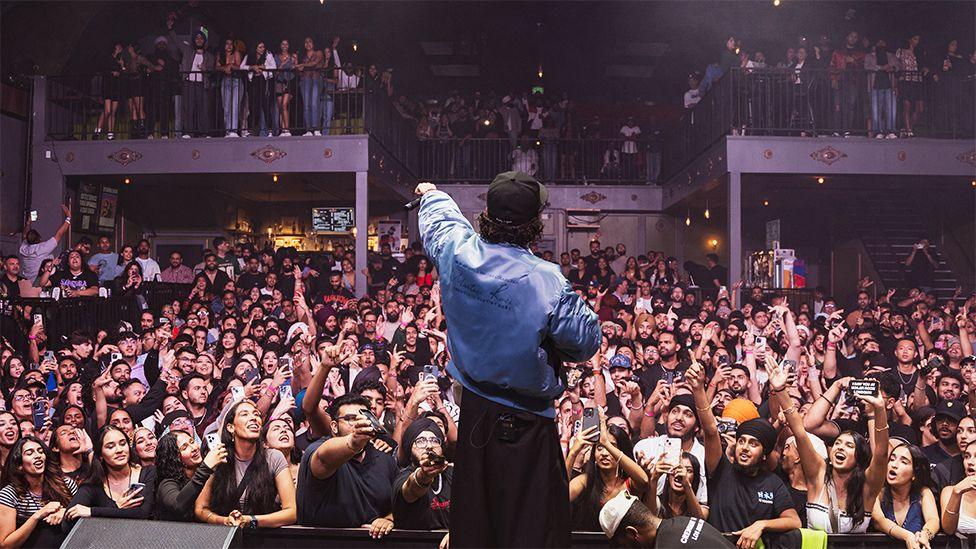 Raf Saperra singing to a large crowd, wearing a blue shirt and black cap, with his left arm outstretched, pointing the black microphone to the crowd. The fans have their hands in the air and are shouting. There are mental stairs and an upper level where fans are also standing.