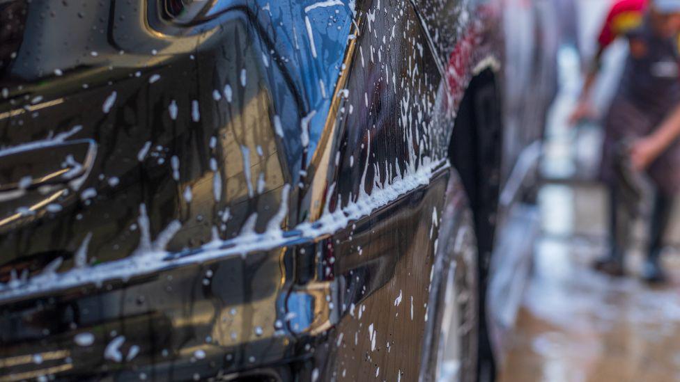 A close up of a black car covered in soap and water. A man can be made out in the background, cleaning a car. 