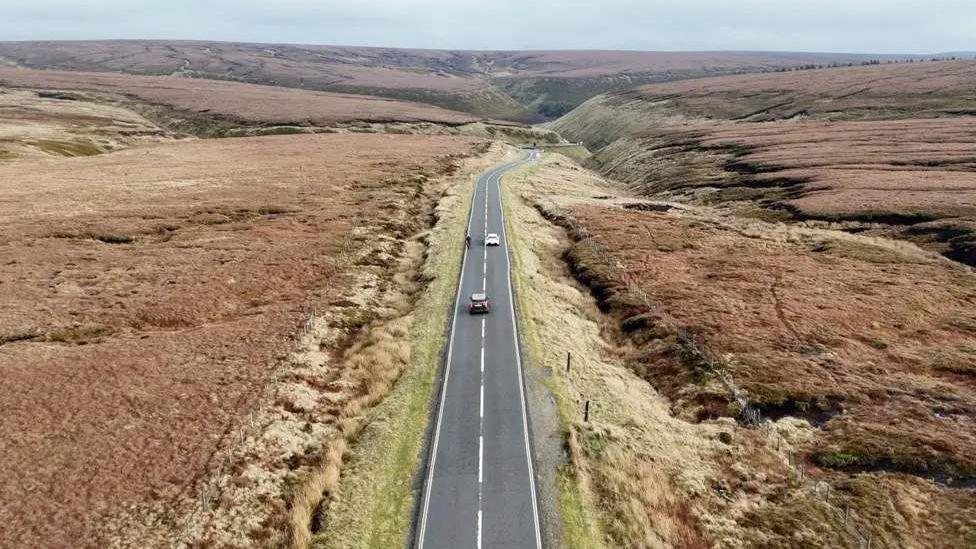 The Snake Pass road winds through open countryside. There are two cars on the road and a cyclist.