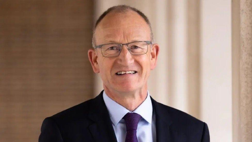 Richard Roberts a man looking directly at the camera and smiling. He is wearing silver rimmed glasses. He is also wearing a black suit jacket, pale purple shirt and purple tie.