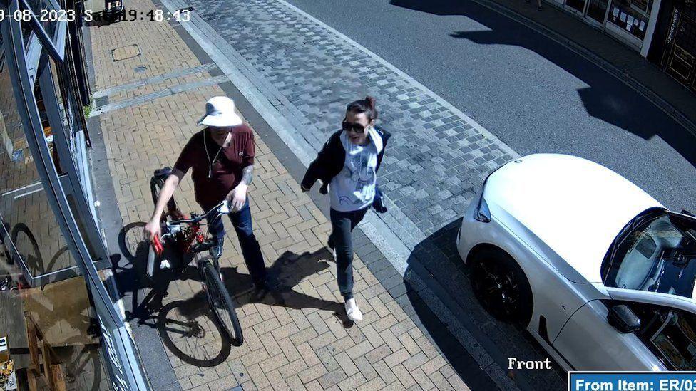 CCTV image of the couple walking along a pavement. Atkins is wheeling a bicycle and is holding a hacksaw