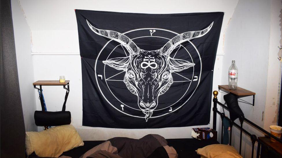 A black banner showing a goat's head in the middle of a pentagram is hung up on a bedroom wall. Beside it is an empty bottle of coca cola and a chocolate box is on the floor