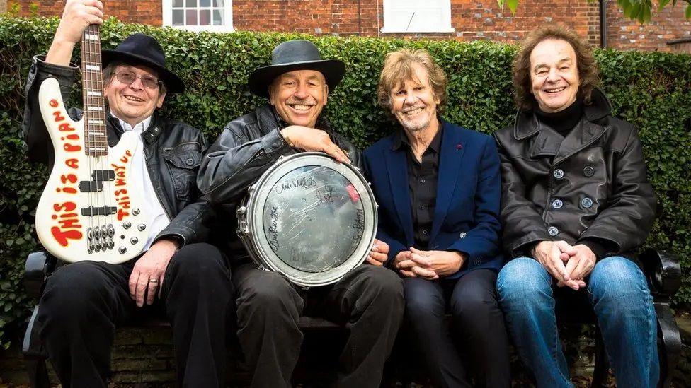 Four members of The Zombies band pictured sitting in St Albans in front of a hedge, two holding their instruments - an electric guitar and a drum. They are smiling cheerfully at the camera.