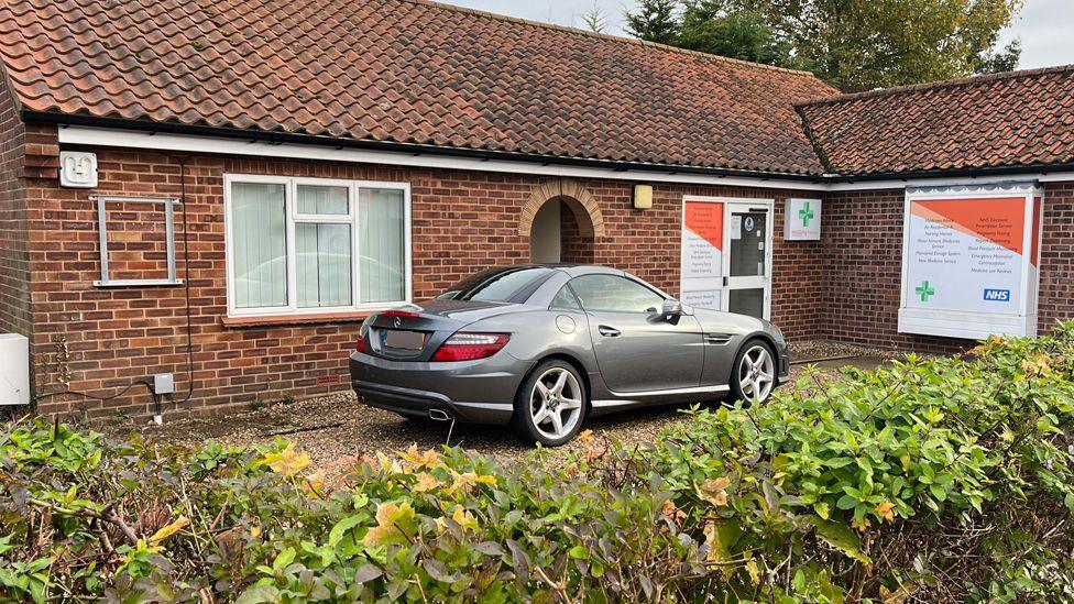 An exterior view of Toftwood Medical Centre. It is a brick building, much more like a house than a modern medical facility. There are signs offering walk-in flu and covid vaccines. 