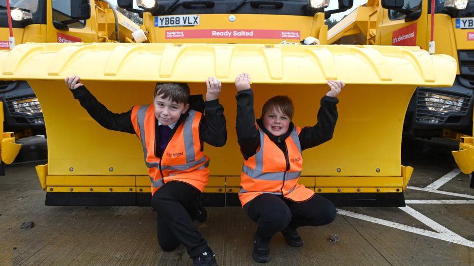 Two school boys wearing hi-vis jackets are crouching under the plough of a large yellow gritter. They have their arms raised and are holding the gritters. A red sign on the gritter announces its name as Ready Salted.