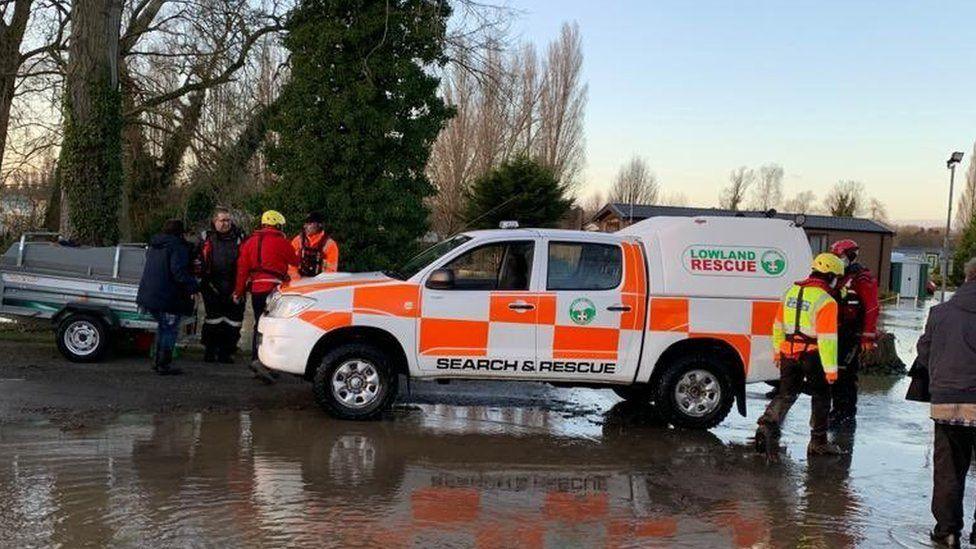 A orange and white van with "Search and Rescue" written on the side is parked up in a large pool of flood water. Several volunteers in orange and red high-visibility jackets, hard hats and hiking boots are walking around. 