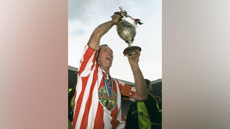 Kevin Ball wearing a red and white football kit and carrying a trophy aloft.