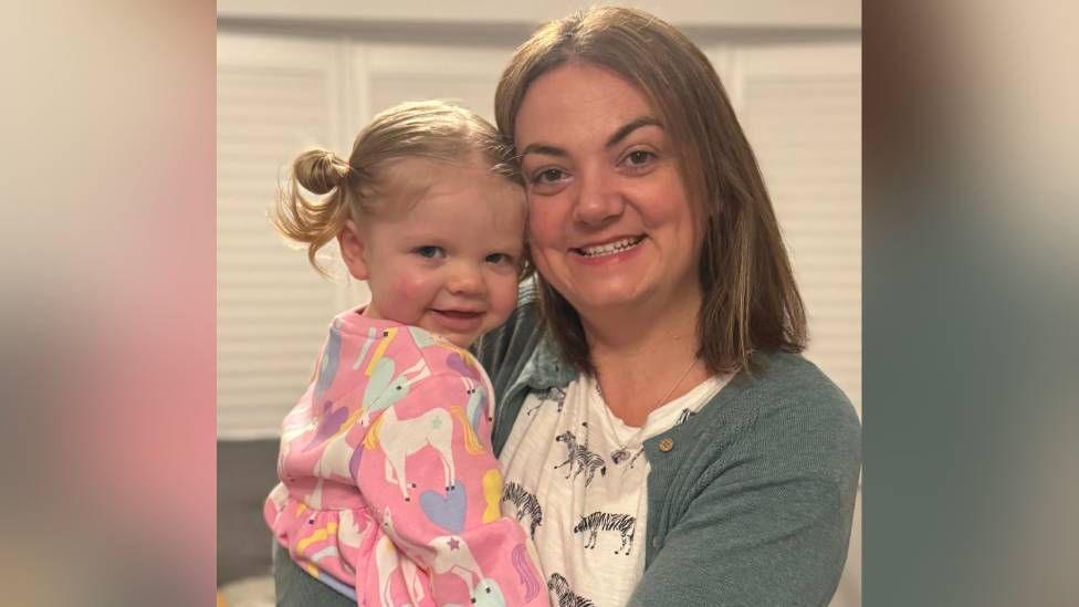 A young girl with blond hair and a pink jumper with horses on is hugged by her mum, who has short brown hair and is wearing a grey/blue cardigan and white t shirt