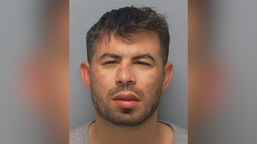 A police mugshot of a young man with short hair and black stubble. His nose is wonky like it's been broken. 