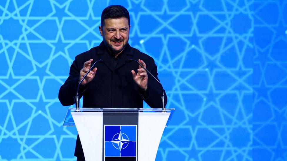 President Volodymyr Zelensky is dressed in a black military jacket and trousers, speaking behind a podium at Nato. He gesticulates whilst he talks into the microphones. Behind him is a blue background with stars on it.