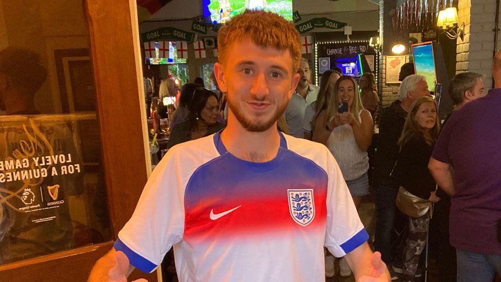 Jacob Williamson standing in the doorway of a crowded pub in Newport Pagnell. He has short fair hair and a short light brown beard. He is wearing an England shirt in red white and blue, starting to smile. Behind him are men and women.
