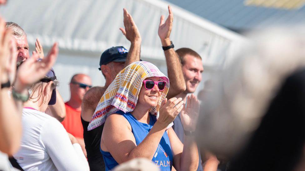 A woman with pink sunglasses and a towel on her head to help cope with the heat claps in the middle of a crowd