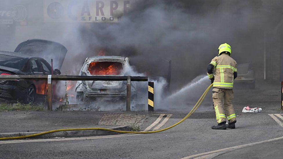 A firefighter uses a hose to tackle blazing in cars on a garage forecourt as plumes of dark grey smoke rise into the air