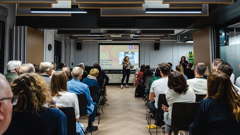 An image taken from the back of a room where a woman is giving a presentation. The powerpoint in the background reads 'Playful Bristol and Youth Work Alliance'. All the seats in the room are full.