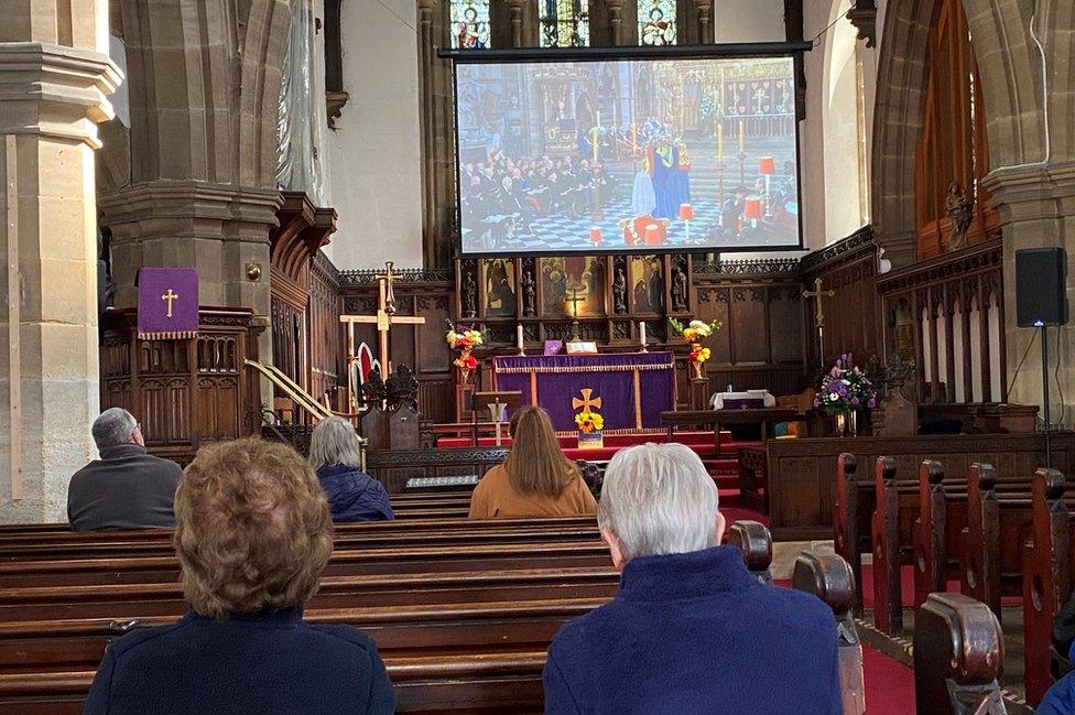 Shot taken from behind of people sat in pews watching the big screen