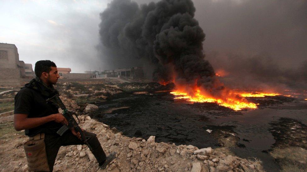 A member of the Iraqi security forces stands with his weapon as fire and smoke rise from oil wells set ablaze by Islamic State militants in Qayyarah (29 August 2016)