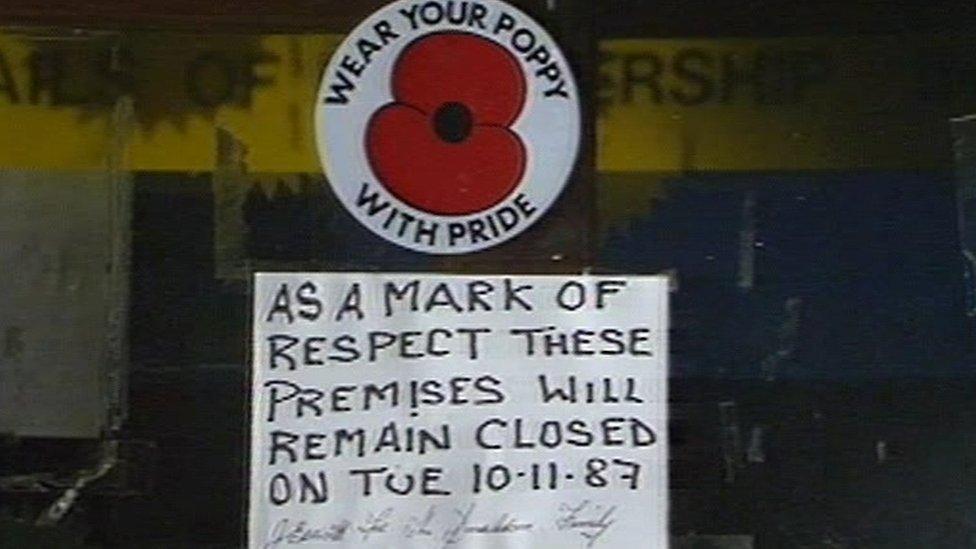 Shop front window from 1987 in Enniskillen with a poppy sticker which says "wear your poppy with pride", placed beneath is a hand written notice which says "as a mark of respect these premises will remain closed on Tue 10.11.87"