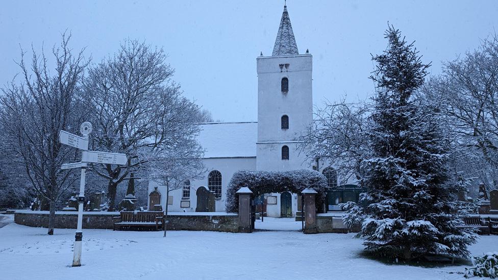 A church covered in snow in Gifford East Lothian. It is partially hidden by bare trees and the ground is also covered in snow. There is a direction sign visible in the foreground.