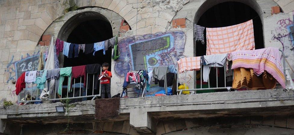 A very young boy stands on a balcony with no barrier or railing at the derelict building, surrounded by washing that has been hung out to dry