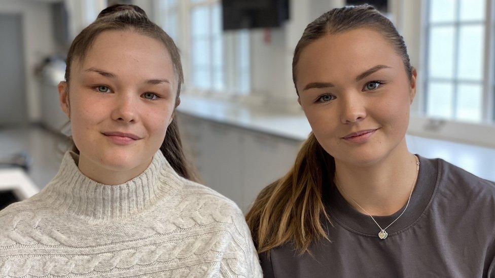 Identical twins sit next to each other in lab. Nancy is on the left in a cream knit sweater and Aimee on the right in a grey t-shirt. Both are smiling