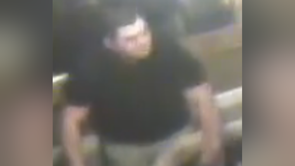 A CCTV still image of a white man wearing a back t-shirt and cream coloured trousers