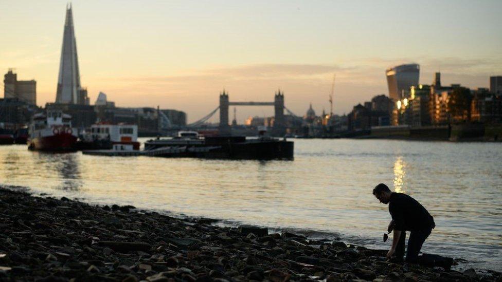 A mudlarker on the Thames foreshore with Tower Bridge, the Shard and the river in the background